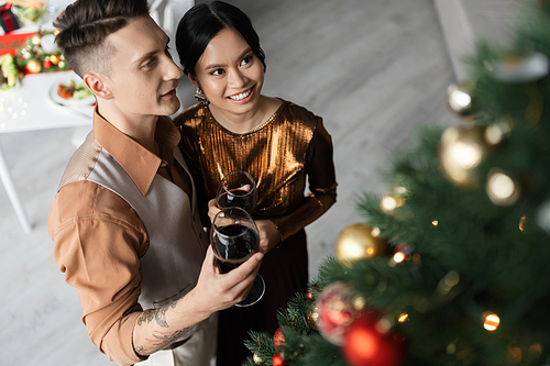 high angle view of happy interracial couple in festive outfits holding glasses of wine near Christmas tree
