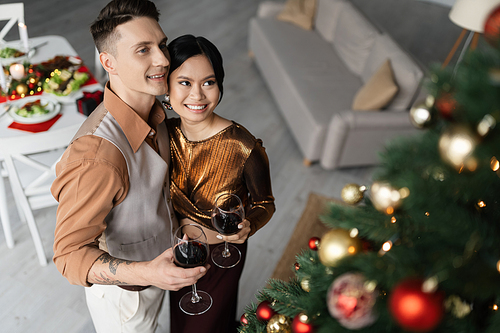high angle view of cheerful interracial couple in festive outfits holding glasses of wine near Christmas tree