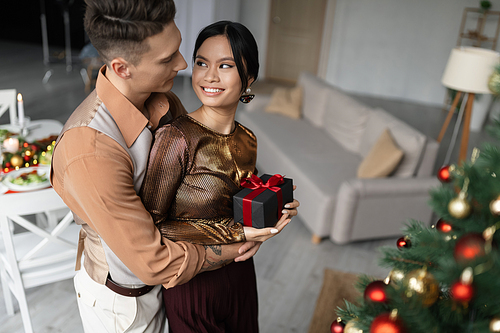 happy and tattooed man hugging asian wife while holding Christmas present