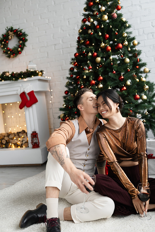 cheerful man kissing cheek of happy asian wife while sitting under Christmas tree