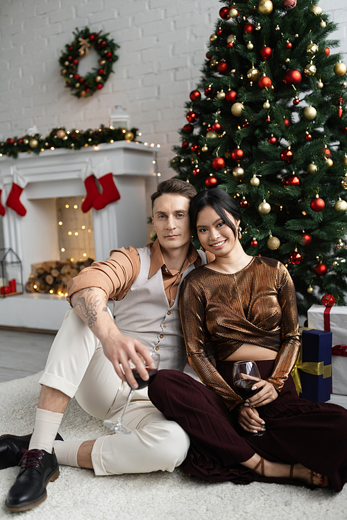 cheerful man and happy asian woman holding glasses of wine while sitting under Christmas tree