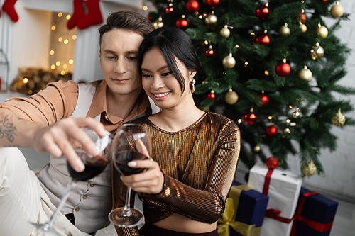 cheerful man and happy asian woman clinking glasses of wine while sitting under Christmas tree