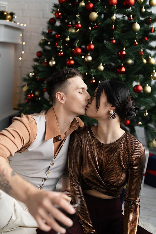 man and asian woman with closed eyes kissing while sitting under Christmas tree