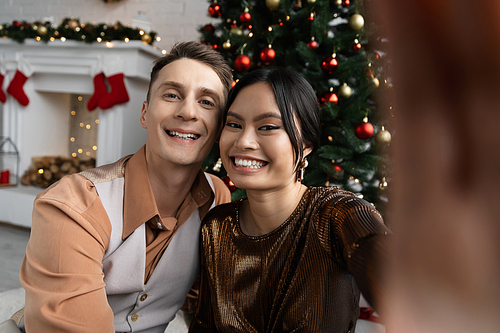 happy interracial couple smiling and looking at camera while sitting under Christmas tree