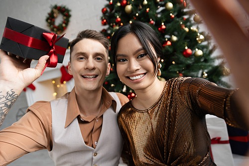 happy tattooed man holding present near asian wife while sitting under Christmas tree
