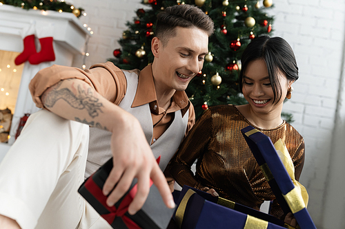 happy interracial couple opening Christmas present while sitting under decorated fir