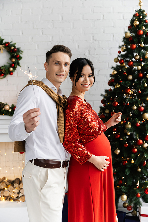 cheerful man holding sparklers with asian pregnant wife near Christmas tree