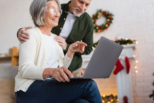 happy middle aged woman in glasses pointing at laptop near happy husband during christmas holidays