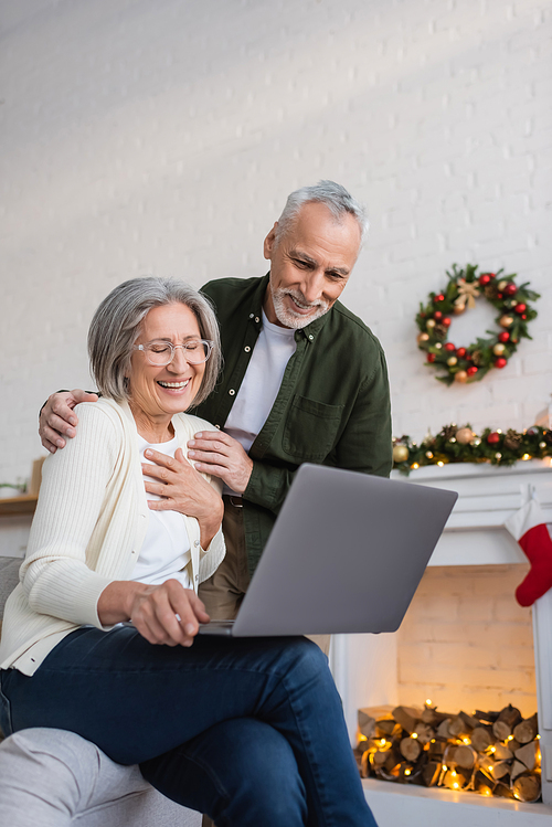 happy middle aged husband and wife laughing near laptop during christmas holidays