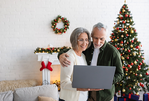 happy middle aged husband and wife looking at laptop near decorated christmas tree