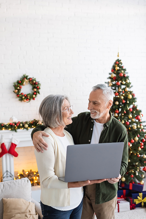 happy middle aged husband hugging wife holding laptop near decorated christmas tree