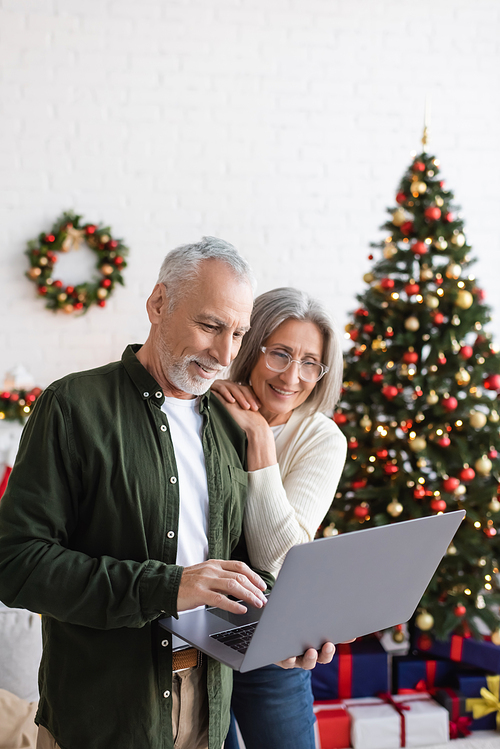 cheerful middle aged husband and wife looking at laptop near decorated christmas tree