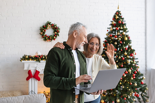 cheerful middle aged man looking at wife waving hand during video chat near decorated christmas tree