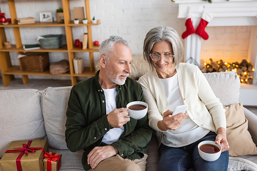 high angle view of mature woman in glasses using smartphone and holding cup near husband during christmas holidays