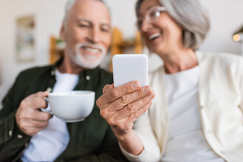 cheerful and mature woman holding smartphone near happy husband with cup of tea