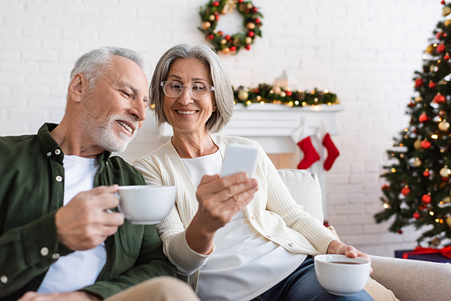 smiling and mature woman in glasses holding smartphone near husband with cup of tea during christmas holidays