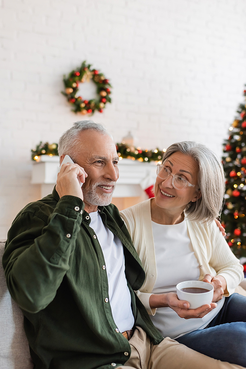 happy middle aged man talking on smartphone near wife with cup of tea during christmas