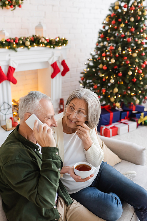 middle aged man talking on smartphone near curious wife with cup of tea during christmas