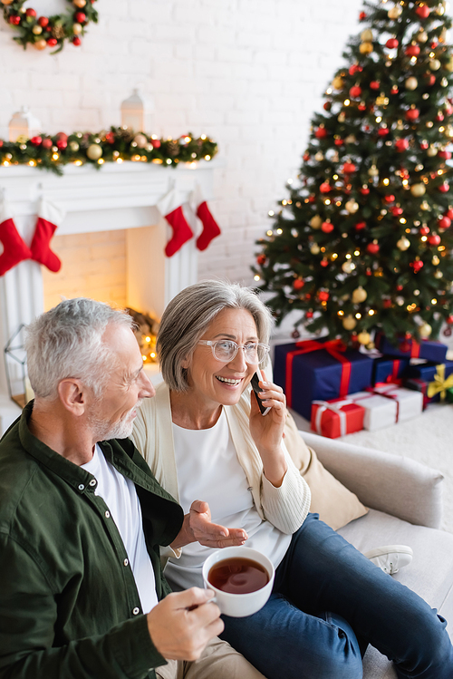 middle aged man holding cup of tea and looking at wife talking on smartphone near christmas tree