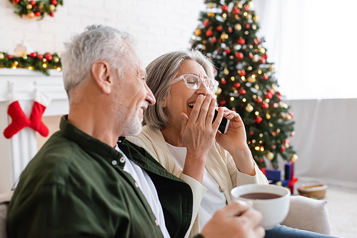 middle aged man holding cup of tea and looking at wife laughing while talking on smartphone near christmas tree