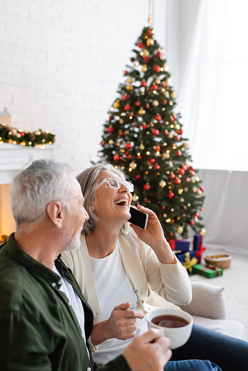 mature man holding cup of tea and looking at wife laughing while talking on smartphone near christmas tree