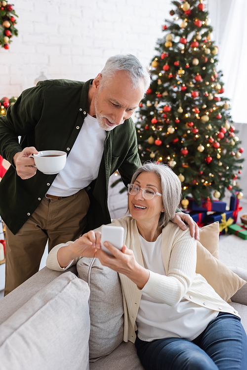mature man holding cup of tea and looking at wife using smartphone near christmas tree