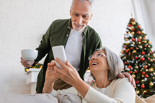 mature man holding cup of tea and looking at smartphone near wife and blurred christmas tree