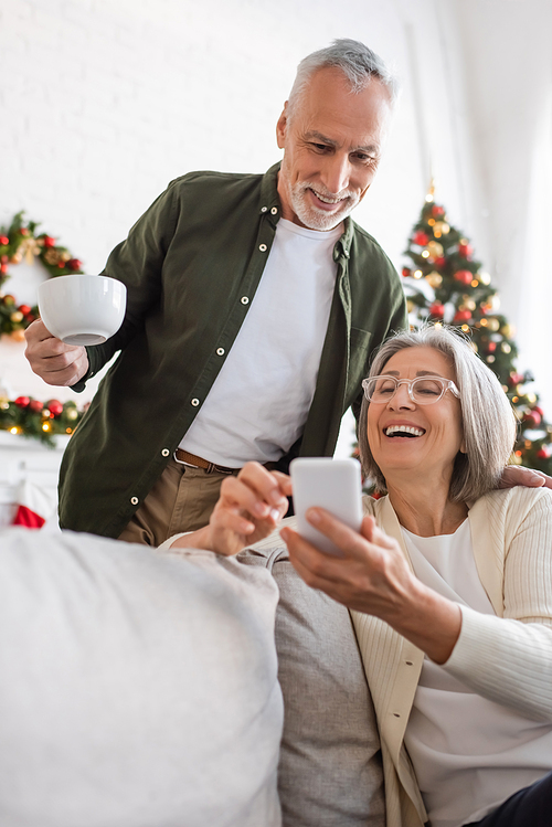 happy mature man holding cup of tea and looking at smartphone near wife and blurred christmas tree