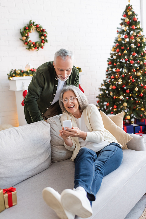happy mature man looking at smartphone near positive wife and christmas tree