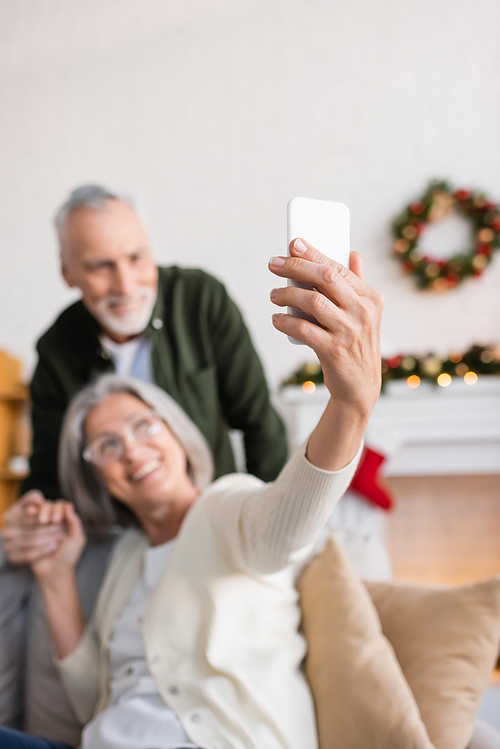 happy mature woman in glasses taking selfie with husband on blurred background