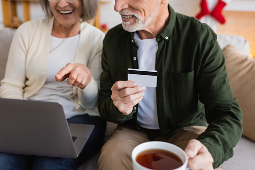 cropped view of mature man holding credit card and cup of tea near wife while having online shopping