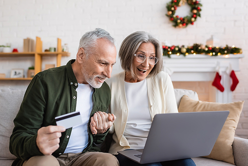 happy mature man holding credit card near excited wife with laptop while having online shopping during christmas