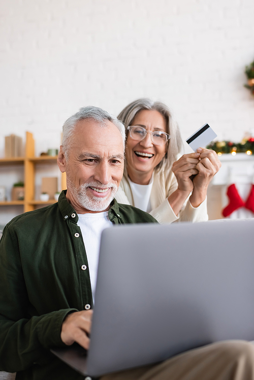 happy mature man using laptop near cheerful wife with credit card during christmas