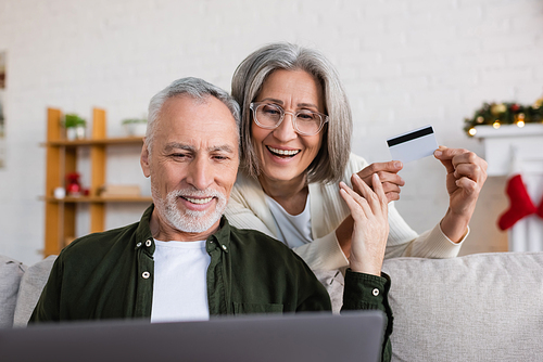 smiling mature man using laptop near cheerful wife with credit card during christmas