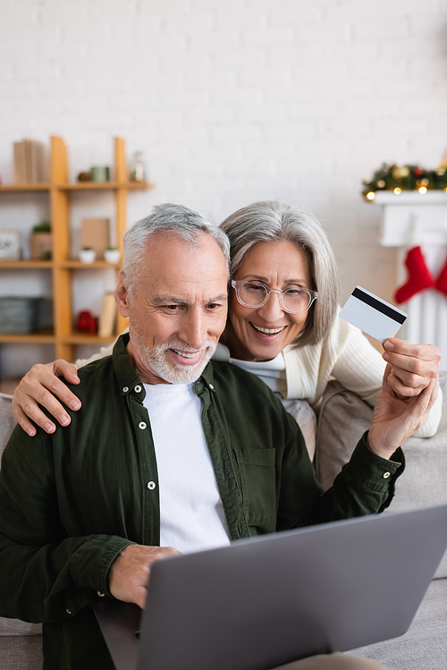 joyful middle aged man using laptop near cheerful wife with credit card during christmas