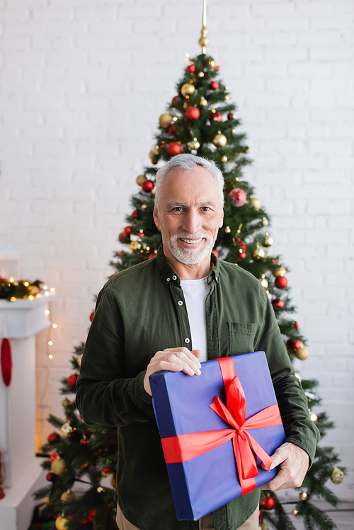 happy and bearded middle aged man holding wrapped present near christmas tree