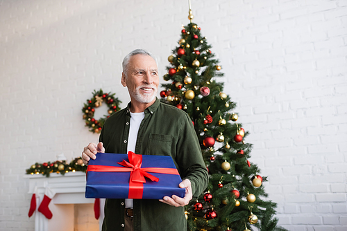 cheerful and bearded middle aged man holding wrapped present near christmas tree
