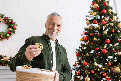 joyful middle aged man with beard holding wicker basket and baubles near christmas tree