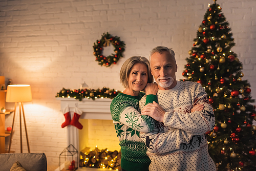 cheerful woman in festive sweater hugging husband near decorated christmas tree in evening