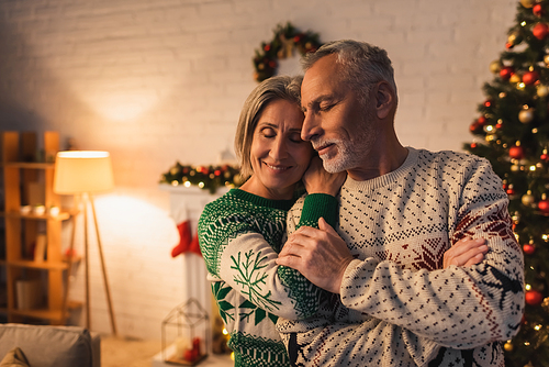 cheerful woman in festive sweater hugging husband with closed eyes near decorated christmas tree in evening