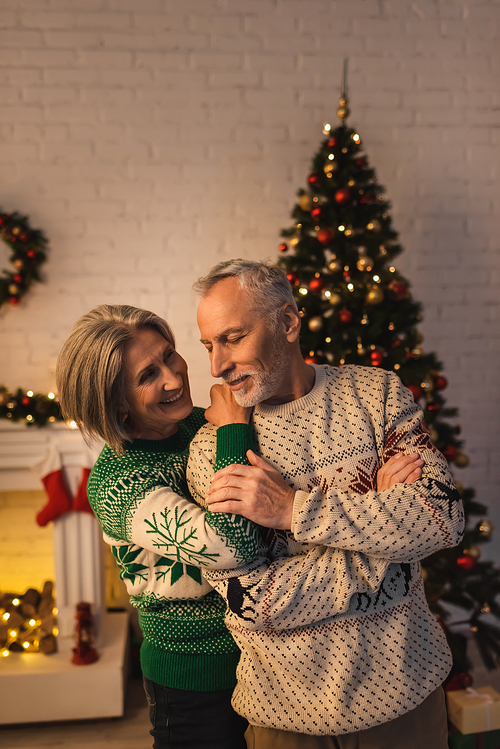 cheerful woman in festive sweater hugging pleased mature husband near decorated christmas tree in evening