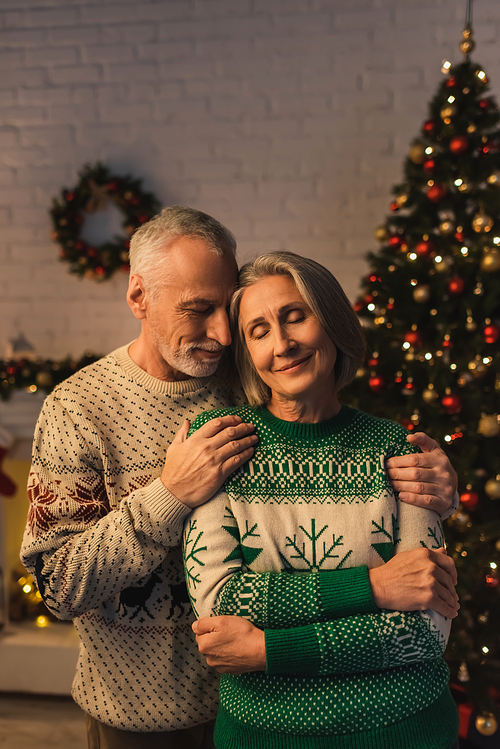 cheerful bearded man in festive sweater hugging pleased mature wife near christmas tree in evening