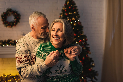 joyful bearded man in festive sweater hugging pleased mature wife near christmas tree in evening