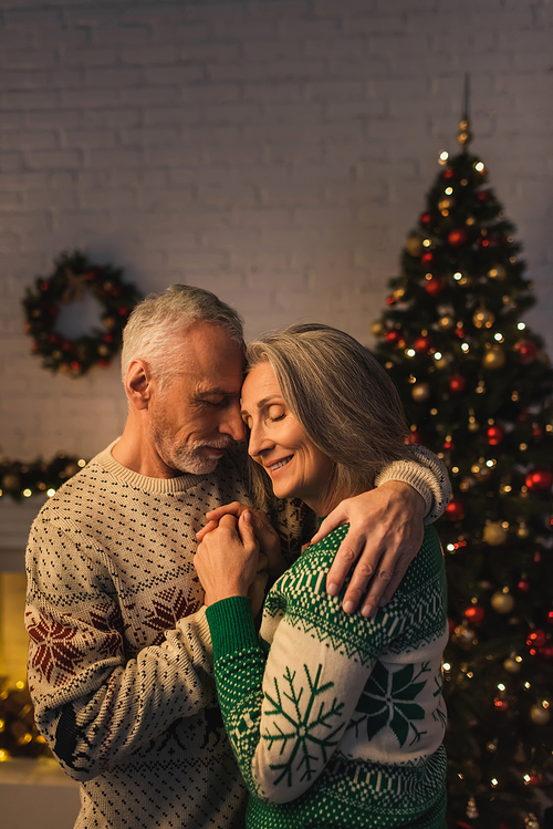 cheerful mature man in festive sweater hugging pleased wife near christmas tree in evening