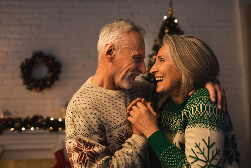 cheerful mature man in festive sweater hugging joyful wife on christmas evening