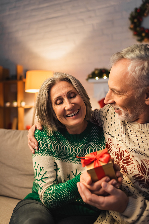 cheerful middle aged woman holding christmas present near husband in sweater