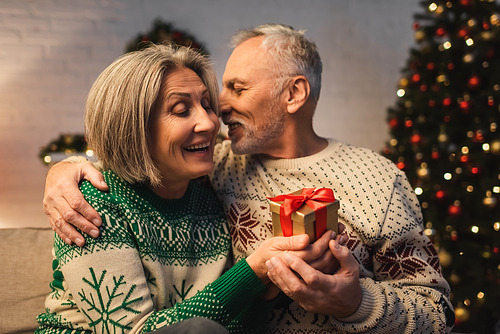 happy middle aged man in sweater hugging smiling wife with christmas present