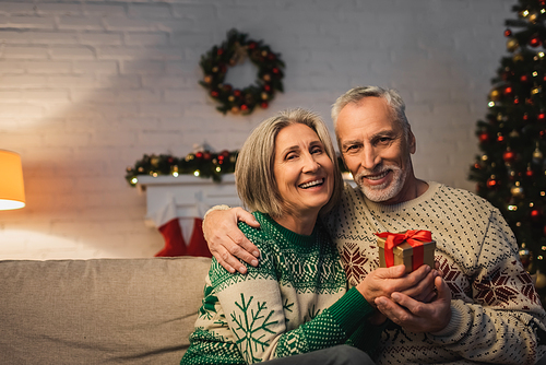 happy middle aged man in sweater hugging cheerful wife with christmas present