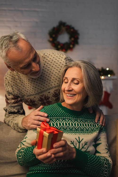 happy middle aged woman holding christmas present near husband in festive sweater