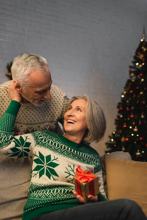 happy middle aged woman holding christmas present and hugging husband in sweater